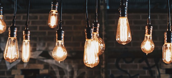 Ten different shaped light bulbs all light up and hanging from the ceiling with a brick wall in the background.