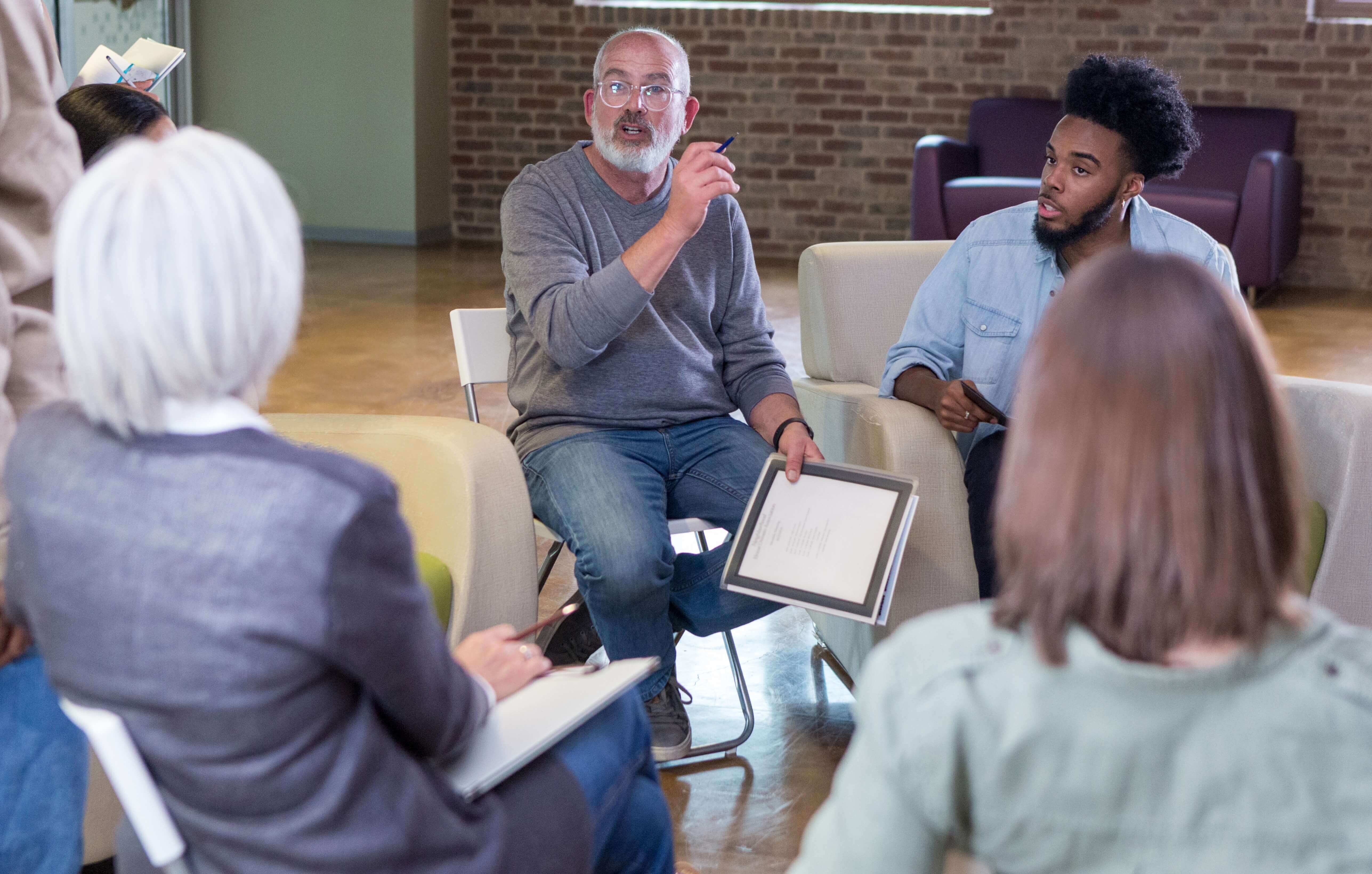 Group of people sitting in a circle. On person is talking and holding papers.